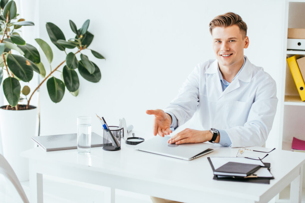 cheerful doctor gesturing while sitting near laptop in clinic